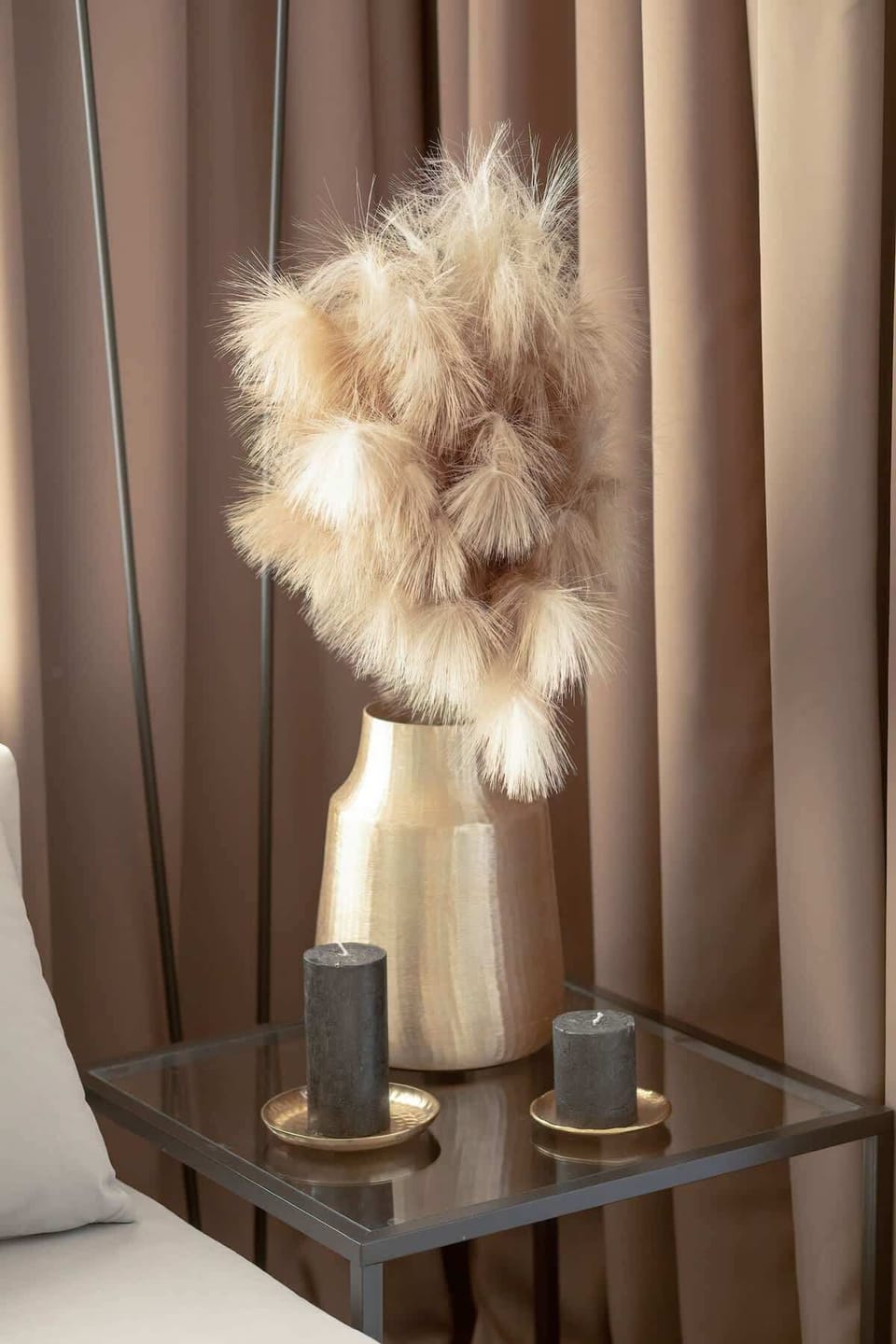 A glass side table with a gold vase filled with fluffy pampas grass and two candles on coasters adds an elegant and cozy touch to the interior.