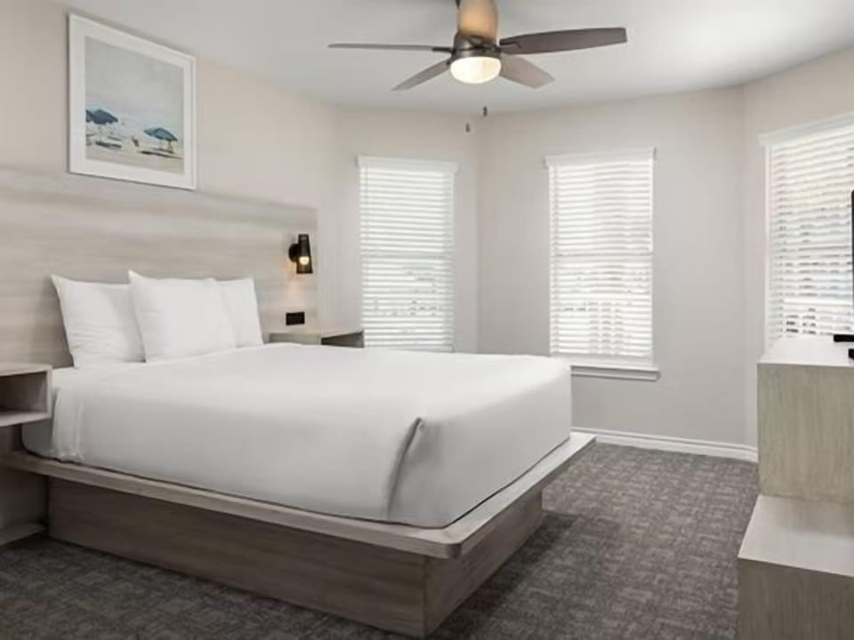 King bed with white bedding, ceiling fan, and large windows with white blinds