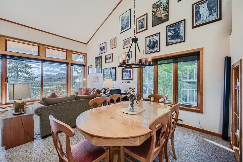 Cozy living and dining area with wooden table, chairs, and couch. Large windows offer scenic views. Walls are adorned with various framed photos and artwork. Ceiling light fixture is a candelabra.