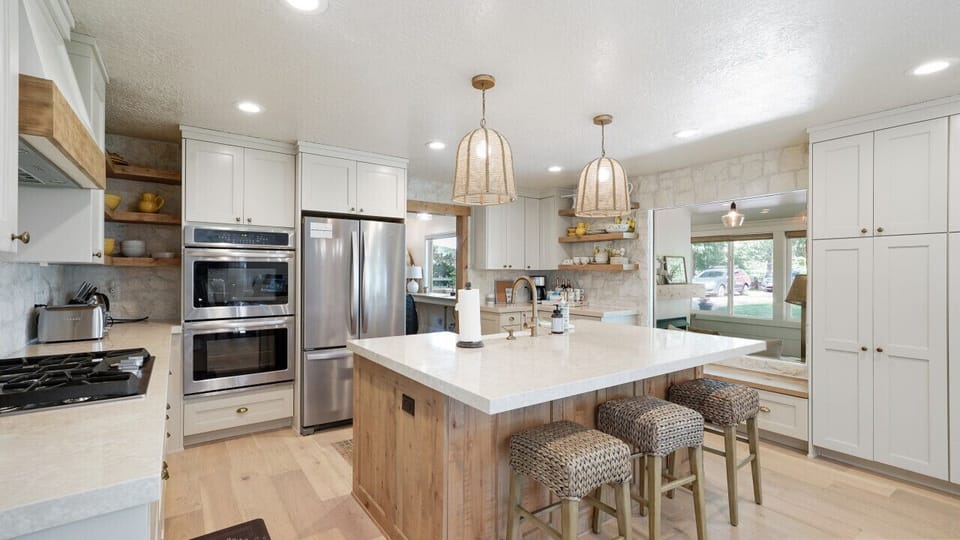 Cook in elegance with a pristine kitchen featuring a white marble countertop, cozy wooden high chair with a knitted cover, modern stove, fridge, and oven, all framed by white-finished cabinets and walls, plus a window that brightens the space.
