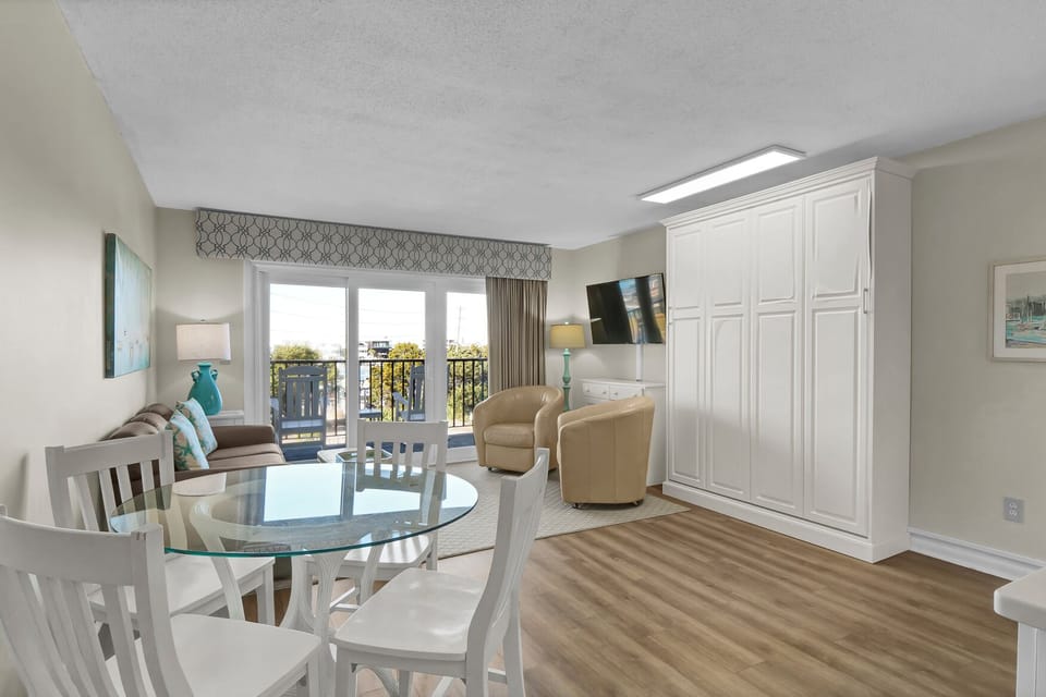 Relax in your spacious suite featuring comfortable living space, dining table, and private balcony access with natural light streaming through.