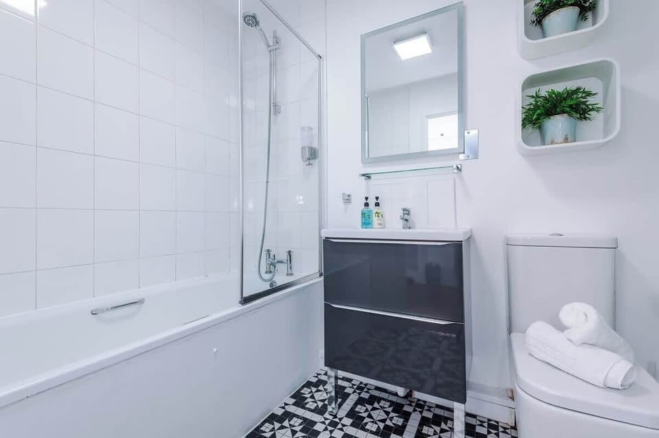 Modern bathroom with a sleek bathtub and glass shower screen, wall-mounted shower, and white tiled walls. Includes a dark vanity with mirror and shelf, stylish black-and-white flooring, and essential toiletries,creating a clean, contemporary space.
