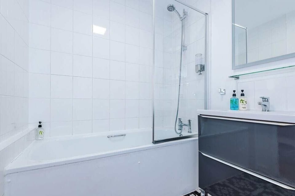 Modern bathroom with a sleek bathtub and glass shower screen, wall-mounted shower, and white tiled walls. Includes a dark vanity with mirror and shelf, stylish black-and-white flooring, and essential toiletries,creating a clean, contemporary space.