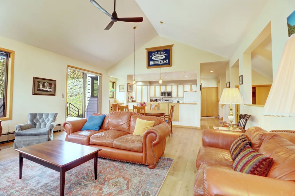 A spacious living room features brown leather sofas, a wooden coffee table, a ceiling fan, large windows, and an adjacent open kitchen with a dining area and barstools.