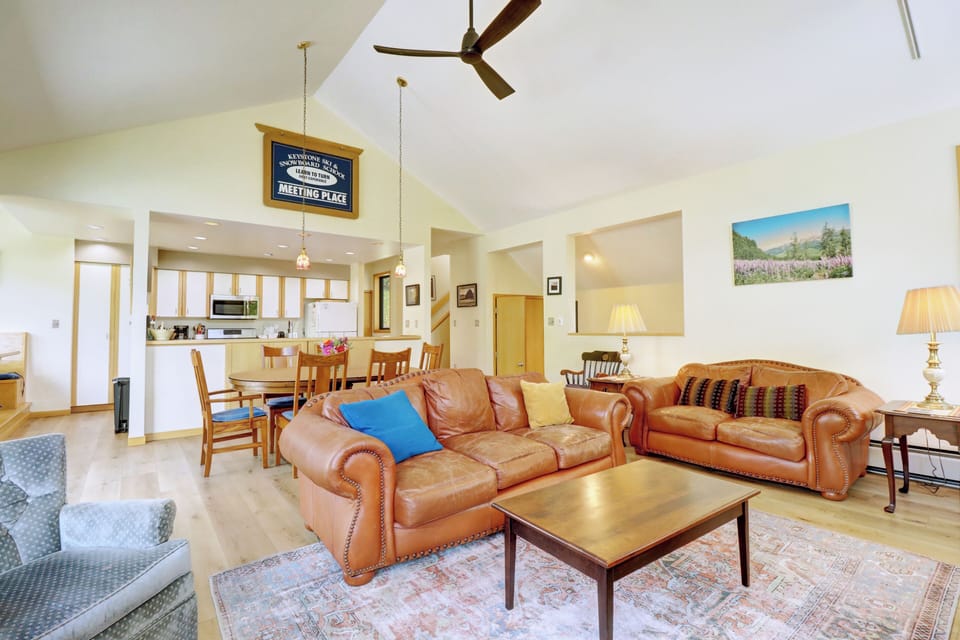 A spacious living room with leather couches, a wooden coffee table, and a ceiling fan. The room connects to a dining area near a modern kitchen. Lamps and framed pictures add to the decor.