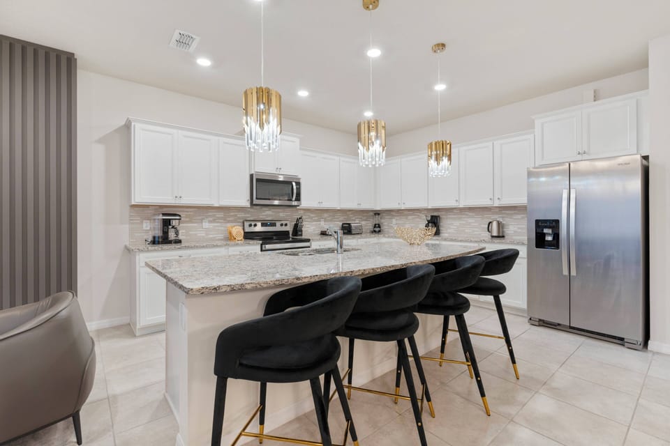 This bright kitchen features sleek white cabinetry with gold accents and a spacious island surrounded by chic black bar stools, perfect for casual dining and offering ample prep space for all your culinary creations.