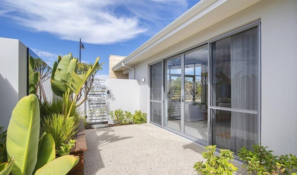 The private outdoor space features a modern paved patio, large sliding glass doors for seamless indoor-outdoor flow, lush tropical plants, a privacy screen, and plenty of natural light under a bright blue sky.
