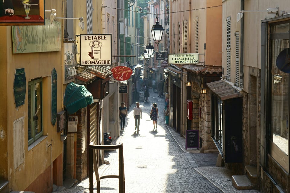 Narrow pedestrian lane in Antibes lined with fromagerie and shop signs, stone facades, and iron lamp posts