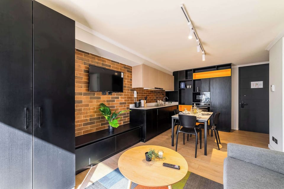 Private studio living area with a four-seat dining table, stylish black kitchen cabinetry, and a warm exposed-brick TV wall.