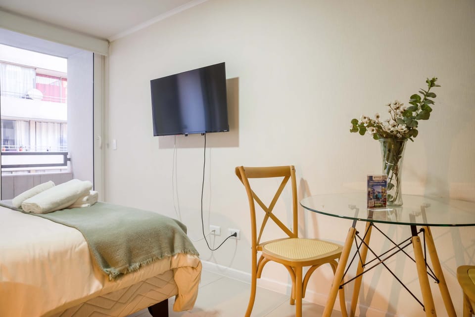 Cozy studio with natural light, featuring a comfy bed, wall-mounted TV, and a charming dining area with a floral touch.