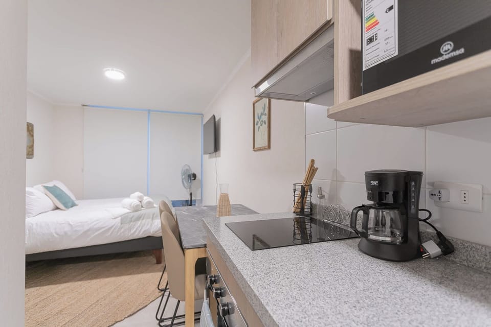 Private kitchen with a black glass cooktop, drip coffee maker, and compact dining counter overlooking the neatly prepared studio sleeping area.
