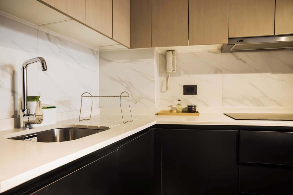Compact studio kitchen with sleek black cabinetry, marble-look backsplash, deep stainless-steel sink, and built-in electric cooktop. (Common Area)