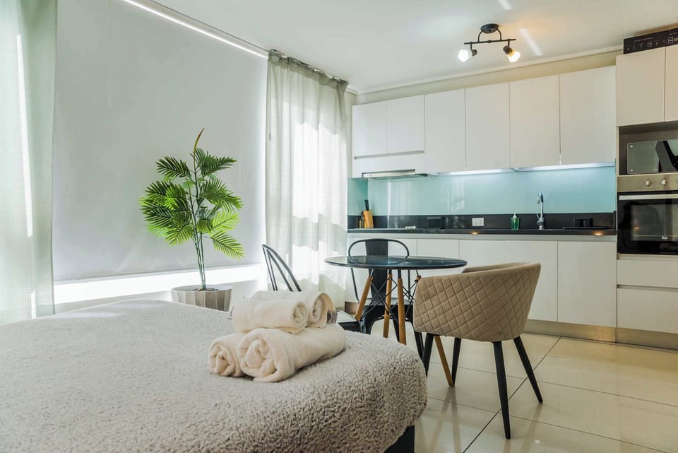 Bright studio kitchen with sleek white cabinetry, a black countertop, and a round dining table beside the neatly made bed.