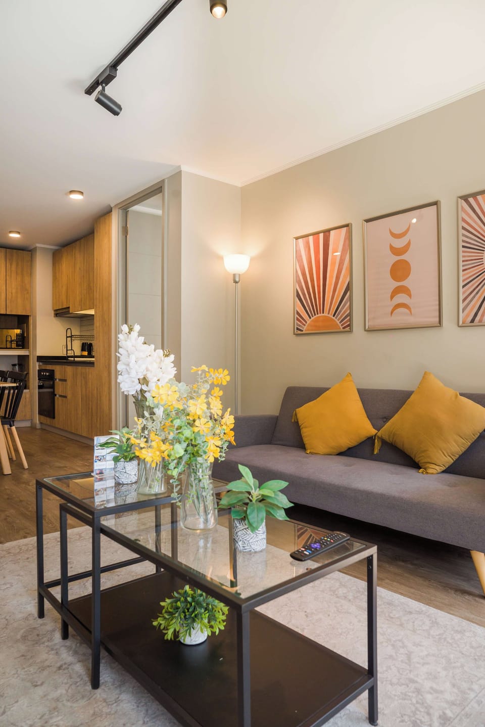 Living room with a gray sectional, mustard cushions, geometric artwork and a compact wood-and-black kitchen.