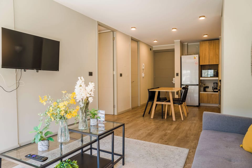Open-plan living room with a wall-mounted TV, gray sofa, and dining area flowing into the compact wood-accented kitchen.