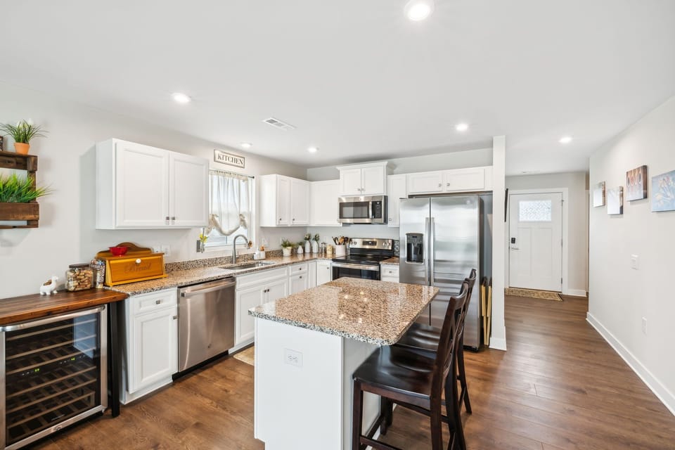 A modern kitchen with white cabinets, granite countertops, stainless steel appliances, a central island with barstools, a wine cooler, and wooden flooring. A door is visible at the far end.