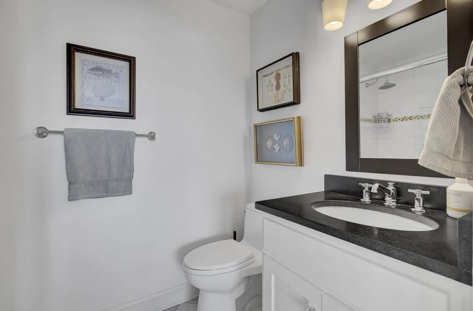 Bathroom with a sink, black countertop, toilet, towel on a rail, and framed pictures on white walls. A round mirror is above the vanity, and a showerhead is visible in the reflection.