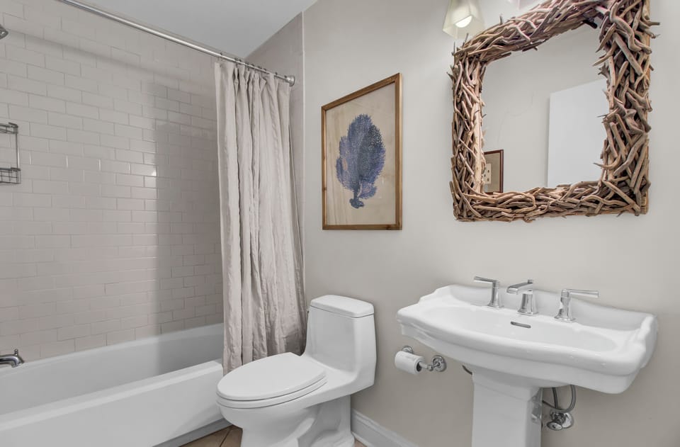 Bathroom with a white bathtub, shower curtain, toilet, and pedestal sink. A driftwood-framed mirror and a framed sea coral print are on the wall.