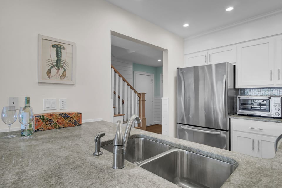 Modern kitchen with a stainless steel fridge, toaster oven, and double sink. A framed lobster print is on the wall. Stairs are visible through an open doorway.