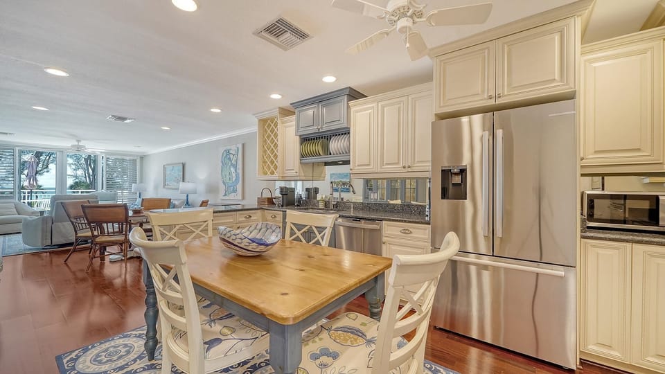 Wood dining table with patterned seat cushions sits atop a blue medallion rug beside cream cabinets and stainless steel fridge.