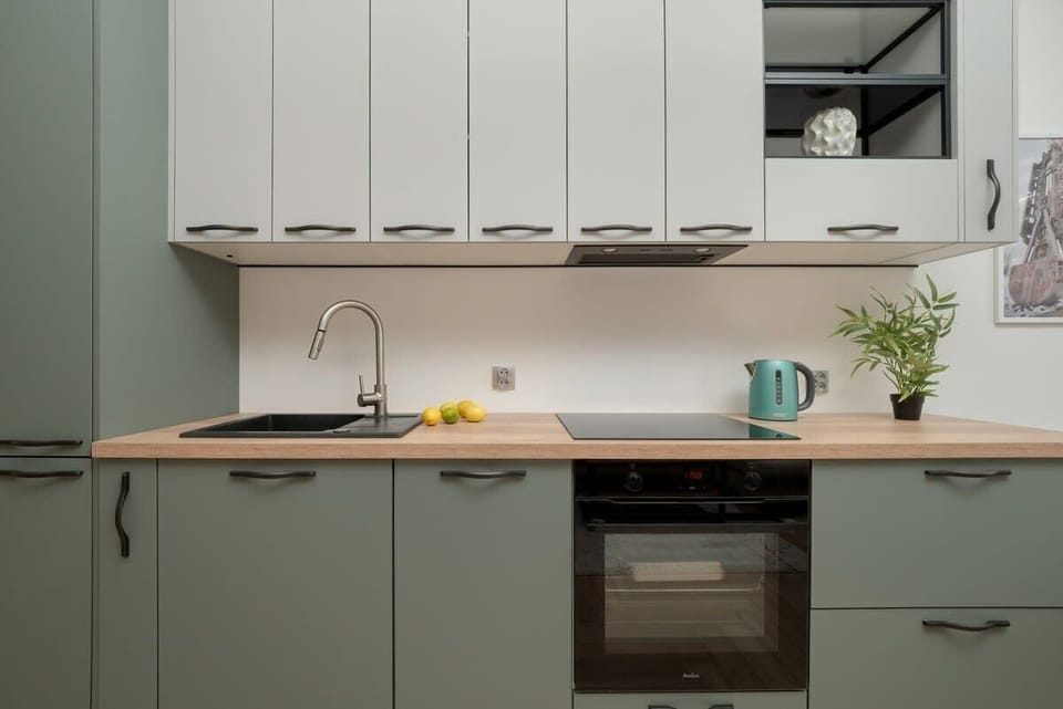 A pastel green and white kitchenette with overhead cabinets, a stovetop, and accessories like mugs and utensils on the counter.
