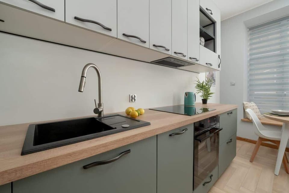 A compact kitchen featuring muted green lower cabinets, a black sink, and light wood countertops with a backsplash in similar tones.