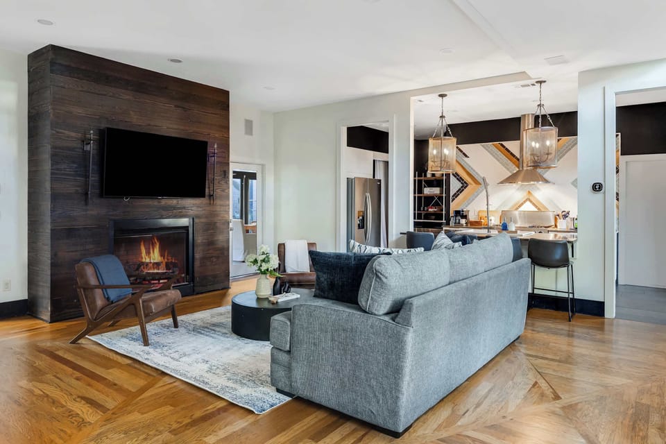 Natural light pours into this beautifully designed living room, highlighting modern furnishings, rich hardwood floors, and warm tones. A smart TV mounted above the gas fireplace makes it easy to stream your favorite shows while relaxing together.