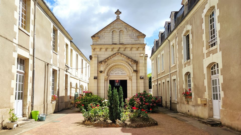 Stone convent facade with arched entrance, flanked by flowering planters and tall hedges