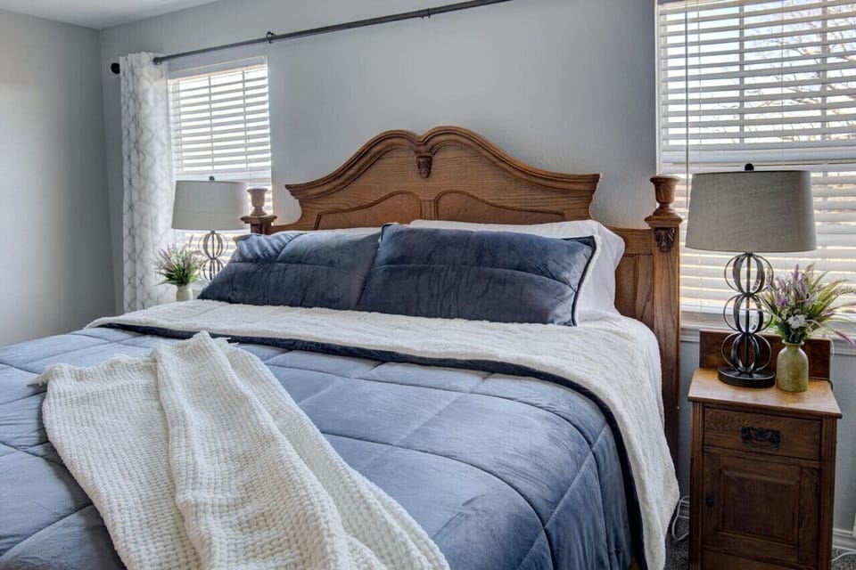Comfortable bedroom scene with a wooden bedframe, soft velvet bedding, wooden side tables with lamps and flower vases, and dual glass windows framed by blinds and curtains