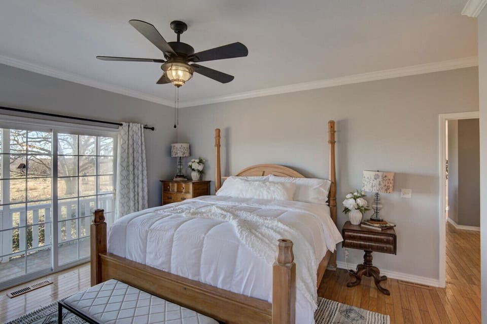 Bedroom 1: A luxurious off-white king suite with wooden ceiling fan, floor, and bedframe. The vintage side table with lamps and flowers adds elegance, and the large glass window with curtains offers a serene view of the farm.