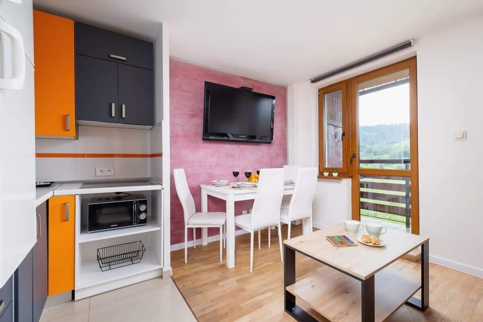 A stylish open-plan dining area with a modern kitchenette, pastel pink walls, and a mounted TV, combining functionality with visual appeal.