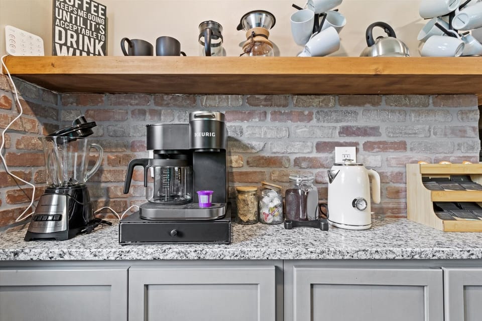 Enhanced coffee bar includes a drip coffee maker, reusable K-Cup, French press, pour over, coffee grounds, creamer and sugar.