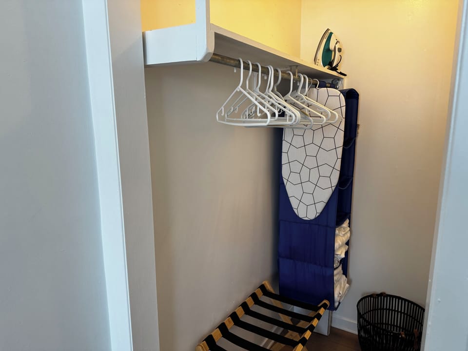 Closet with iron, ironing board, suitcase stand, hangers, and storage.