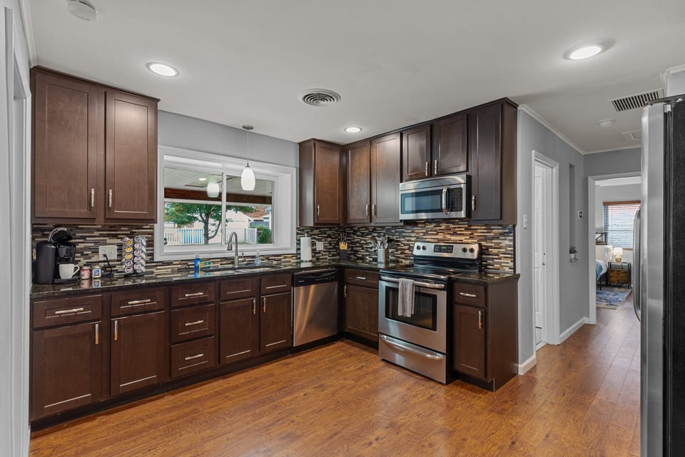 Reverse kitchen view showing the sink, dishwasher, cooking appliances, cabinetry, and hallway beyond.