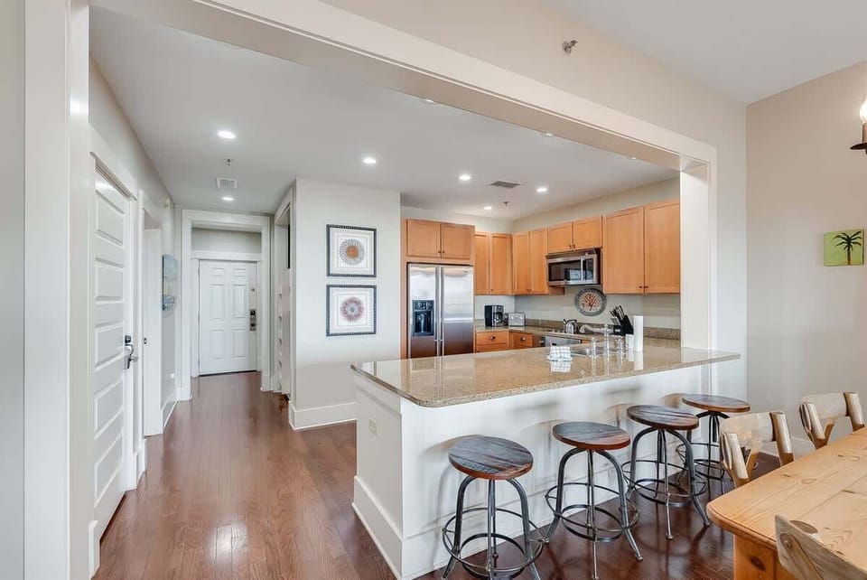 - Spacious granite island offers plenty of space and seating with four rustic barstools - Open concept design seamlessly connects kitchen, hallway, & dining areas - Framed wall art, hardwood floors, and warm lighting create a cozy and welcoming vibe