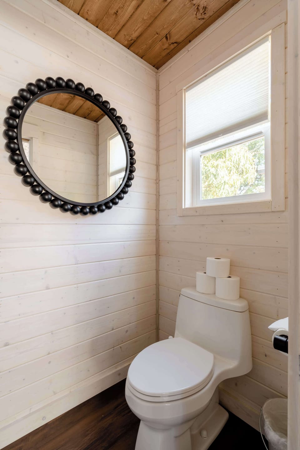 Toilet beneath a round mirror beside a window.