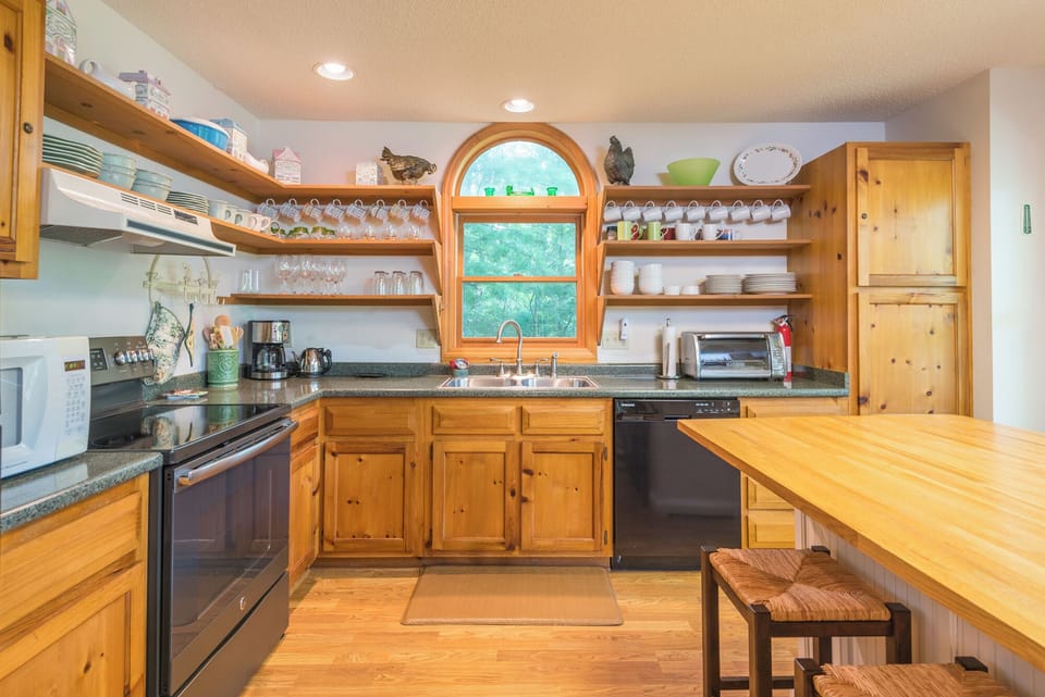 Bright kitchenette-style kitchen boasts stainless appliances, ample counter space, open shelving, and a charming arch-top window overlooking the woods.