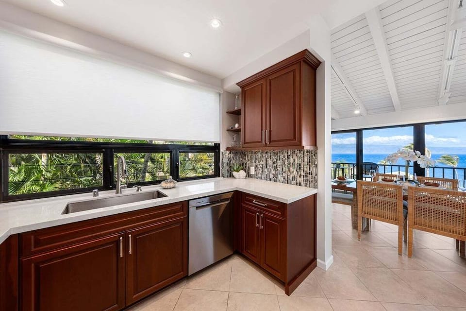 Sunlit kitchen with granite counters, ample cabinetry, and wide windows overlooking lush greenery and glimpses of the ocean beyond.
