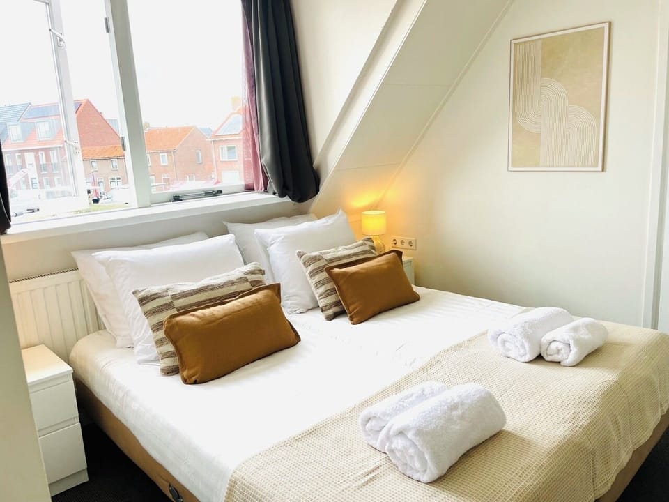 This image showcases a bright and inviting bedroom featuring a neatly made double bed with crisp white linens, beige and brown accent pillows, and a matching throw blanket. The room is illuminated by natural light from a window with a charming view of red-roofed houses, complemented by soft artificial lighting from a bedside lamp. A framed minimalist artwork adorns the wall, ad...