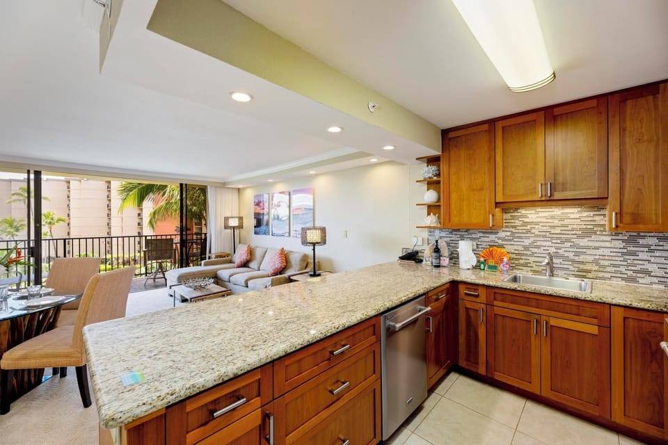This bright and spacious open-concept kitchen and living area features granite countertops, rich wood cabinetry, and seamless access to a private lanai overlooking the tropical resort grounds.