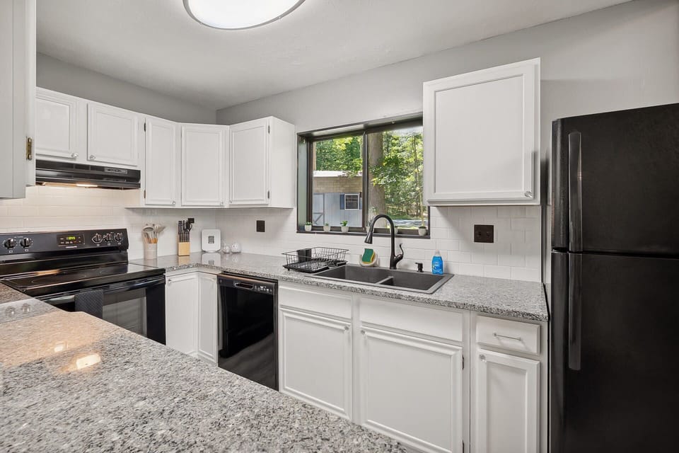Modern kitchen with granite countertops and black appliances overlooking a wooded backyard.