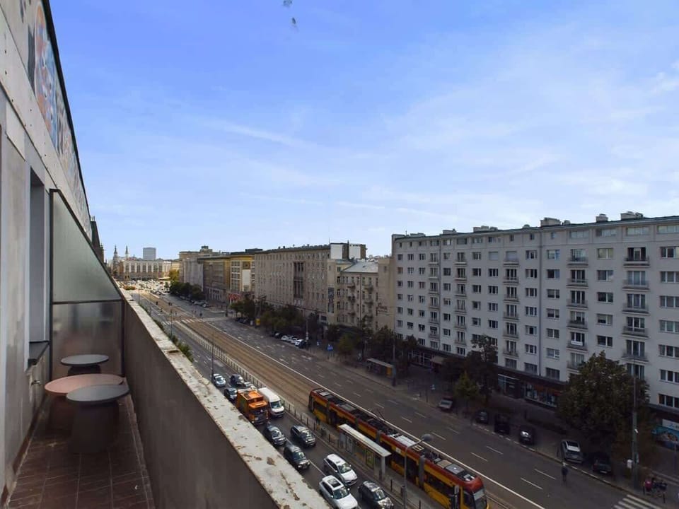 Balcony view overlooking modern residential buildings, blue sky, and tree-lined street.