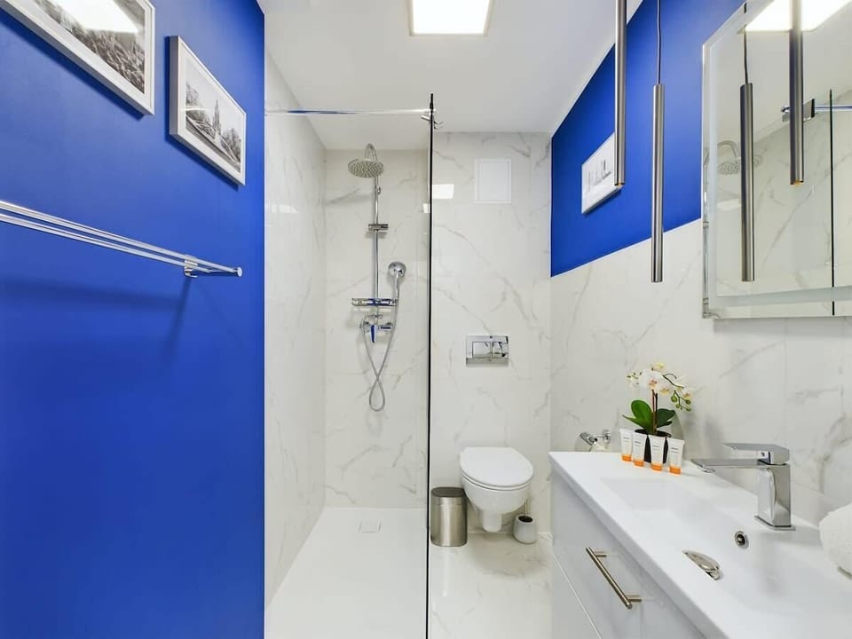 A clean, modern bathroom with white tiles, a walk-in shower, and blue cabinets. A well-lit mirror adds functionality to the space