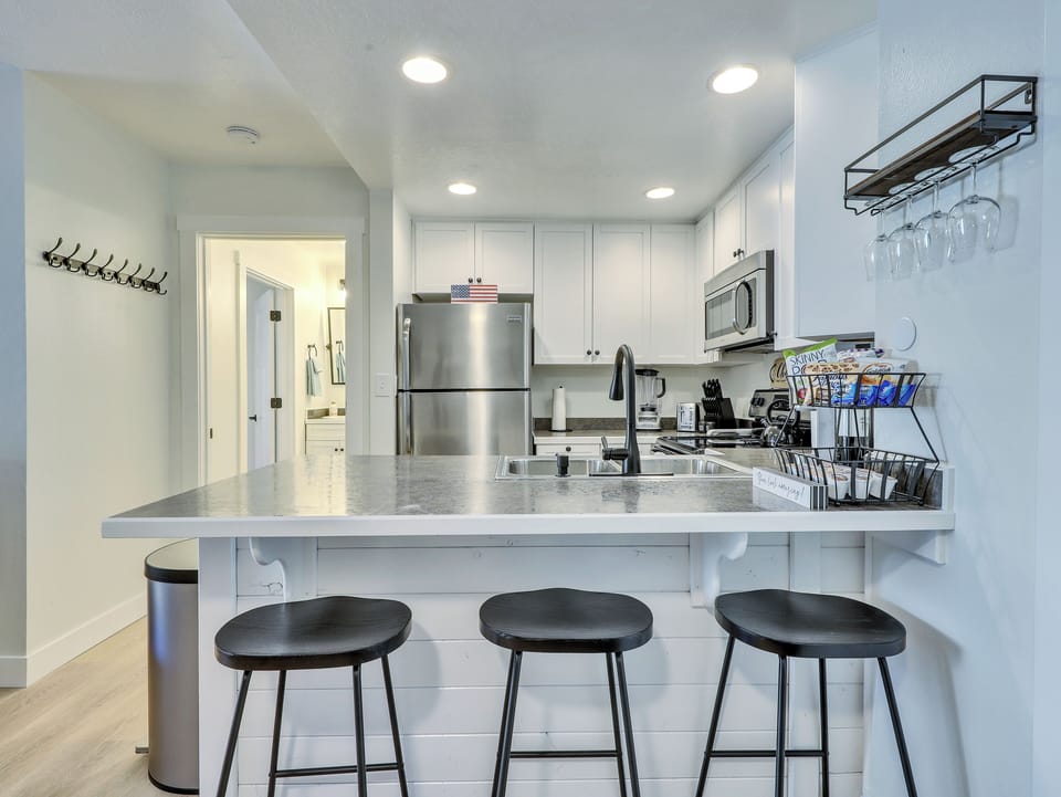 View of the Kitchen Including Microwave, Oven, Stove, Refrigerator, Stainless Steel Sink, Blender, Kettle, Crock Pot, Knife Block, Counter Seating & 3 Stools, Wine Glasses, Cooking Spices, Keurig & Drip Coffee Machine