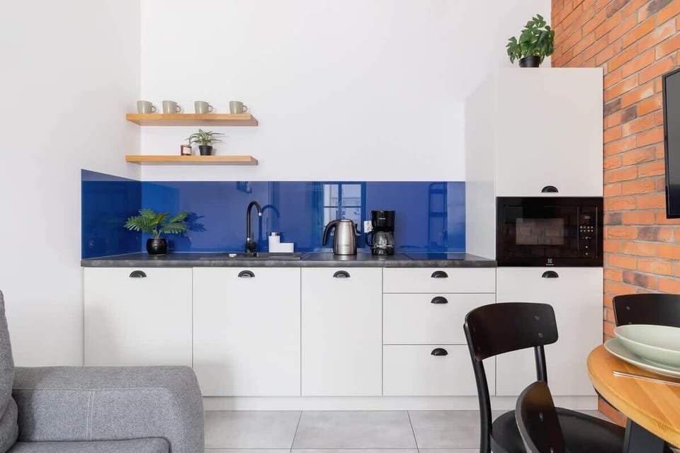 A modern kitchen area with white cabinetry and a pop of blue on the backsplash. A small gray chair and shelf with plants add charm.