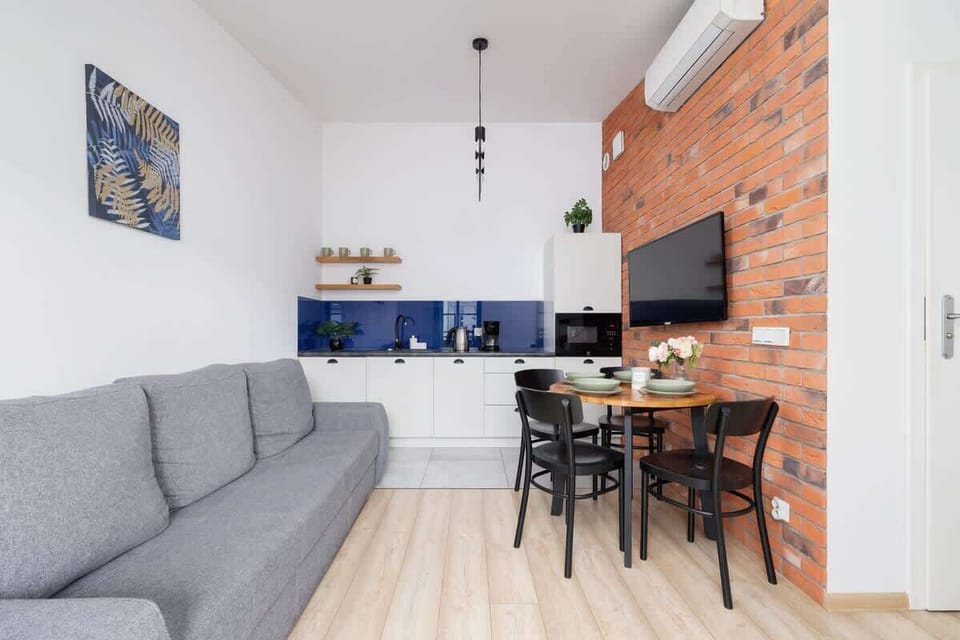 A welcoming living room and kitchen area with a brick wall, gray sofa, and compact kitchen setup. Warm lighting adds to the homey vibe.