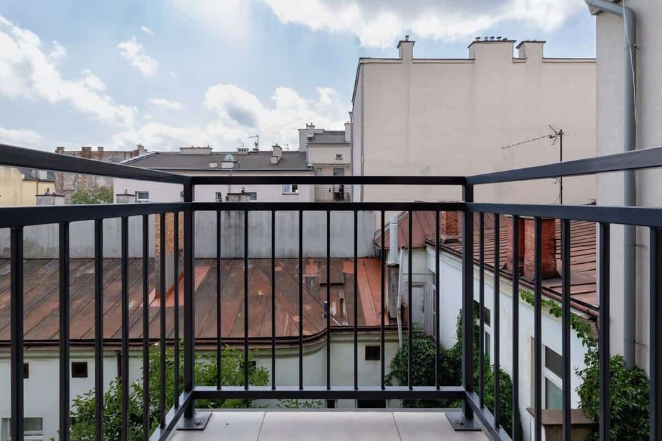 A city view from a balcony showing rooftops and buildings. Metal railings frame the peaceful urban scene.