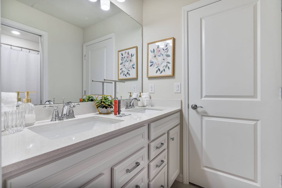This bright and airy bathroom boasts a spacious double vanity with sleek white cabinetry and modern fixtures. The charming floral artwork and large mirror create an inviting space, ideal for a refreshing start to your day.