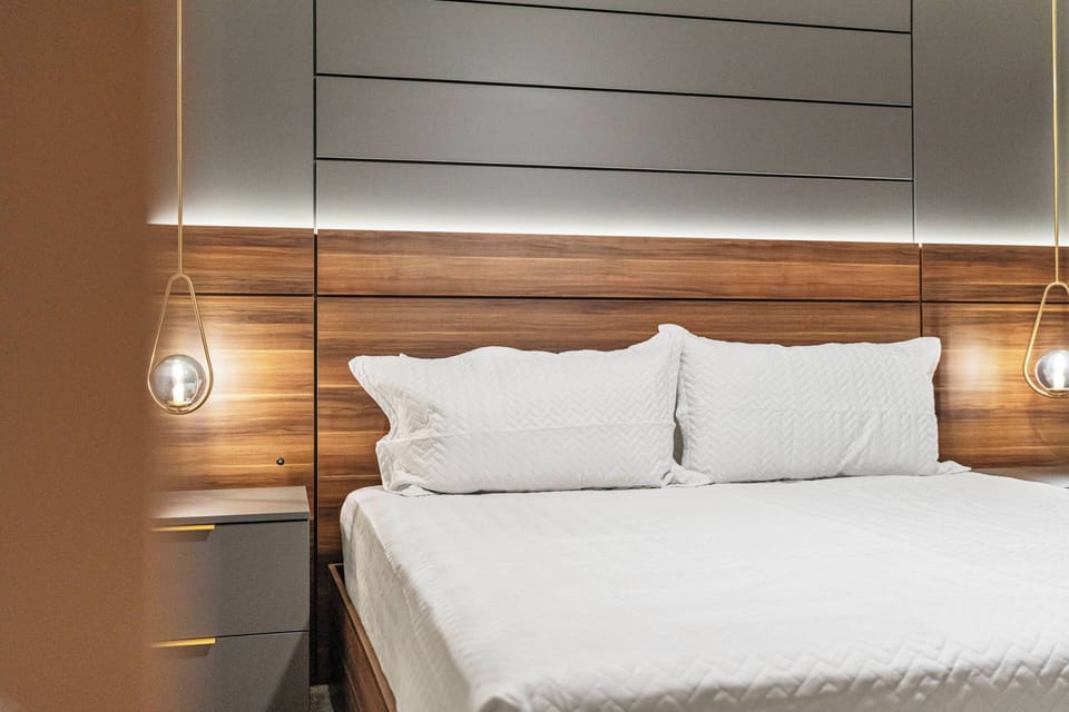 Experience a restful night in this beautifully designed bedroom with a plush bed, crisp white linens, and warm wooden accents, all complemented by soft lighting from stylish pendant fixtures for your ultimate relaxation.