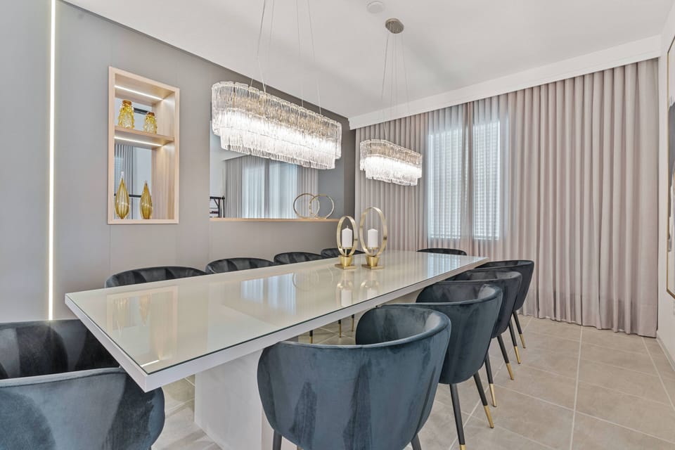 Enjoy a dining area that blends modern design and comfort, featuring a sleek glass table and plush dark chairs. Crystal chandeliers add warmth, enhancing the elegant decor and soft, neutral tones for a welcoming atmosphere.